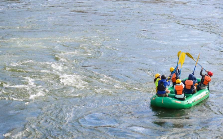 A group of students wearing safety gear sit in a raft and raise their paddles into the air.